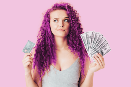 woman with curly pink hair looking unsure holding a credit card and money in her hands