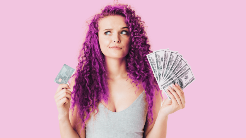 woman with curly pink hair looking unsure holding a credit card and money in her hands