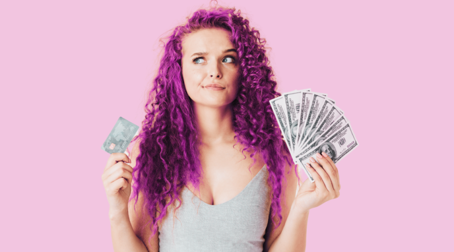 woman with curly pink hair looking unsure holding a credit card and money in her hands